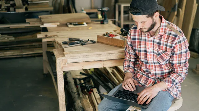 Tischler sitzt in seiner Werkstatt und arbeitet am Laptop