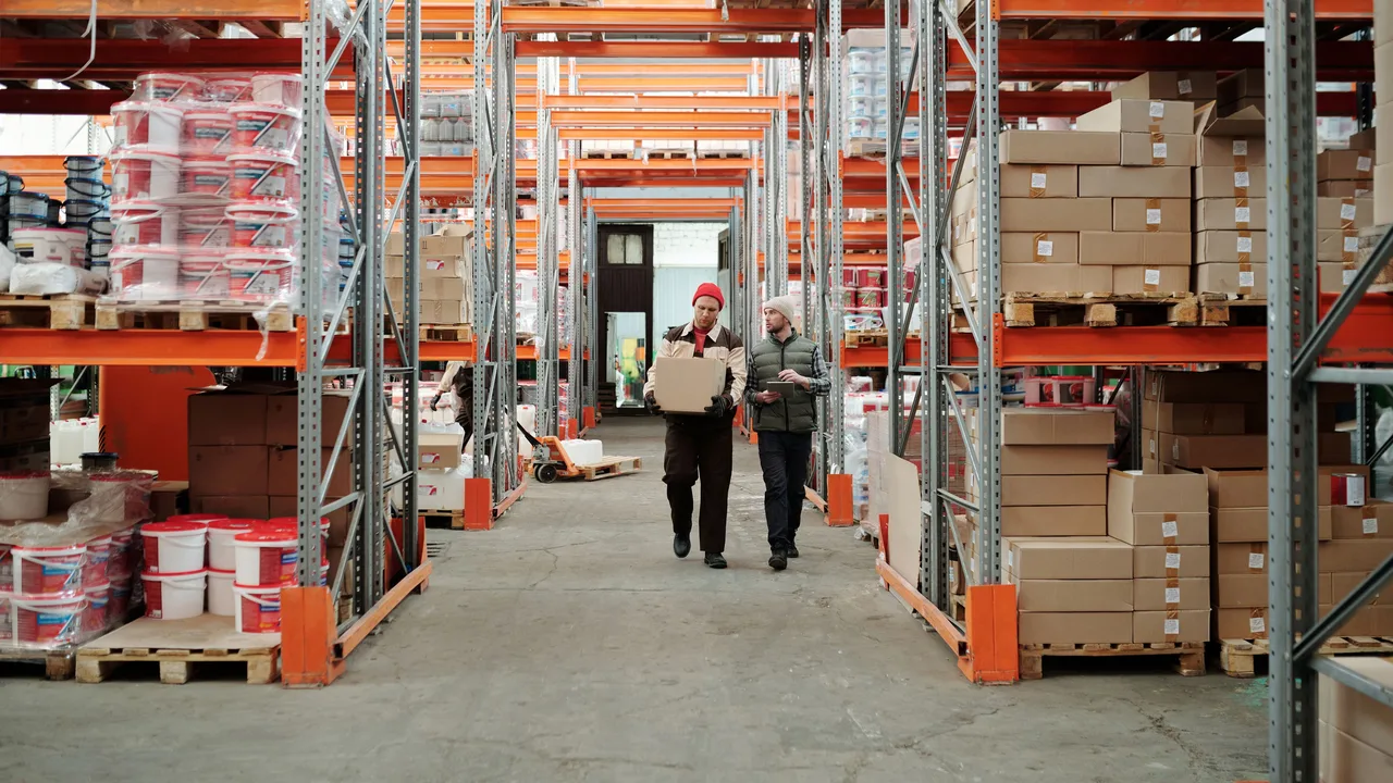 Zwei Mitarbeiter tragen ein Paket durch den Gang eines Logistiklagers